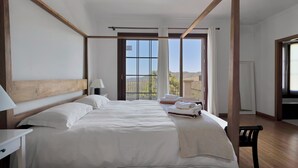 Un grand lit à baldaquin avec du linge de lit blanc, une tête de lit en bois et une vue sur l’extérieur grâce à de grandes fenêtres.