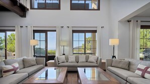 Un salon spacieux avec un grand canapé d’angle, deux tables basses en verre et une vue sur l’extérieur grâce à de grandes fenêtres.