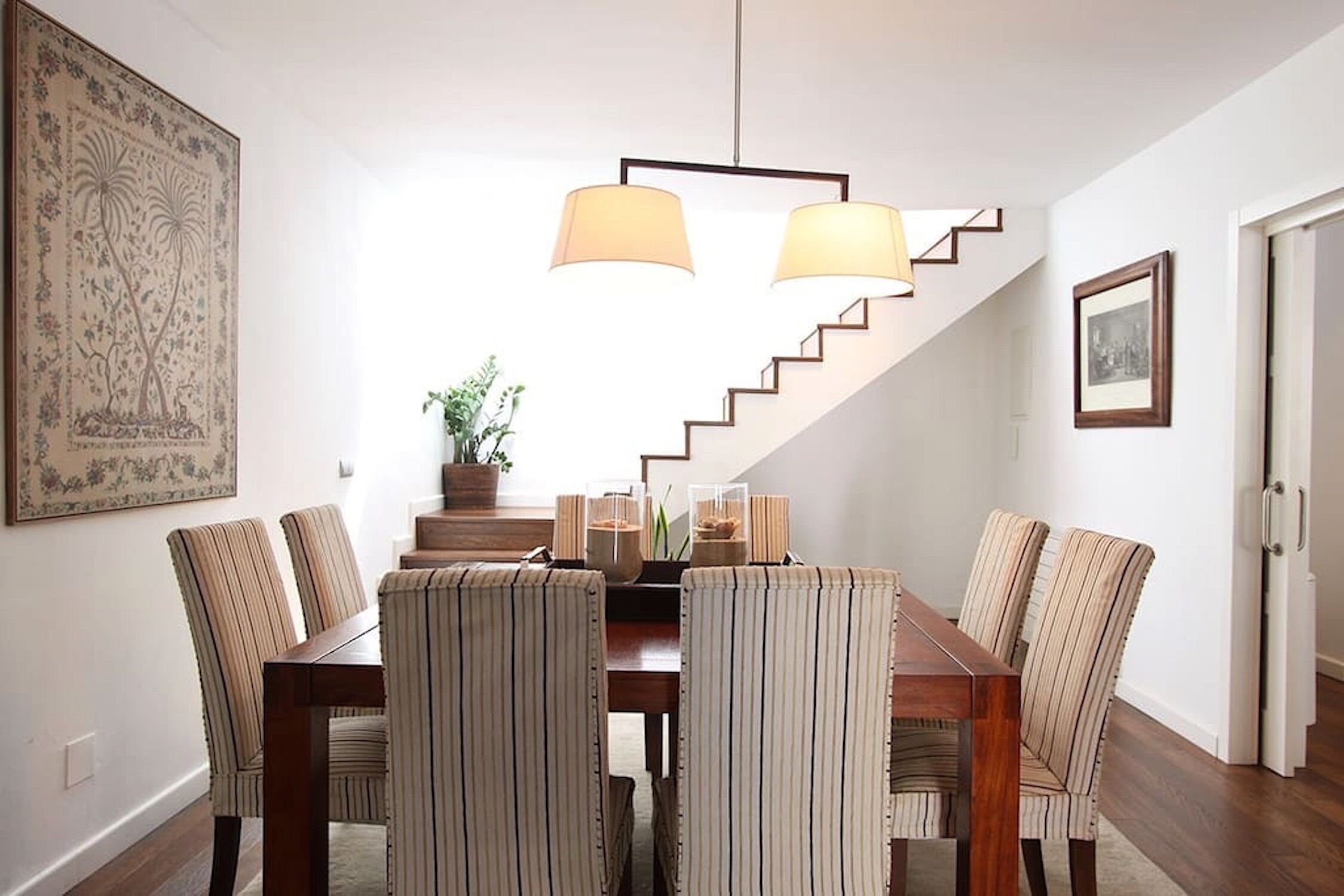 A dining area with a wooden table and striped chairs, a staircase, and a hanging light fixture.