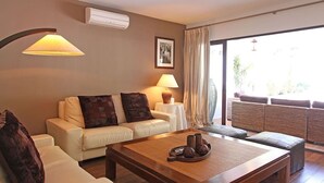 Een woonkamer met een bank, een salontafel en een lamp. Er is een raam met gordijnen en uitzicht op groen.