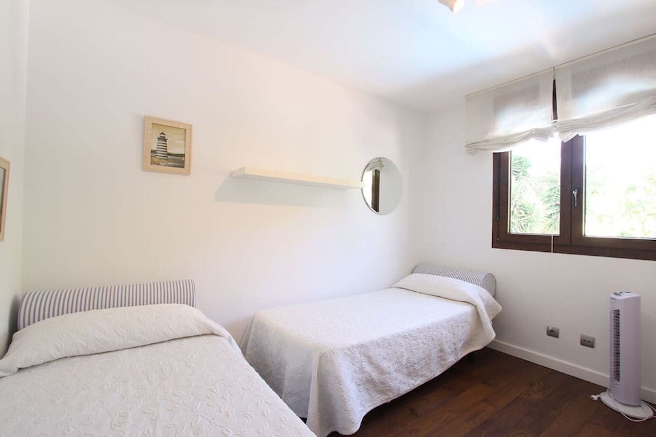Una habitación con dos camas individuales, una mesita de noche, un espejo instalado en la pared, una ventana con cortinas y un radiador.