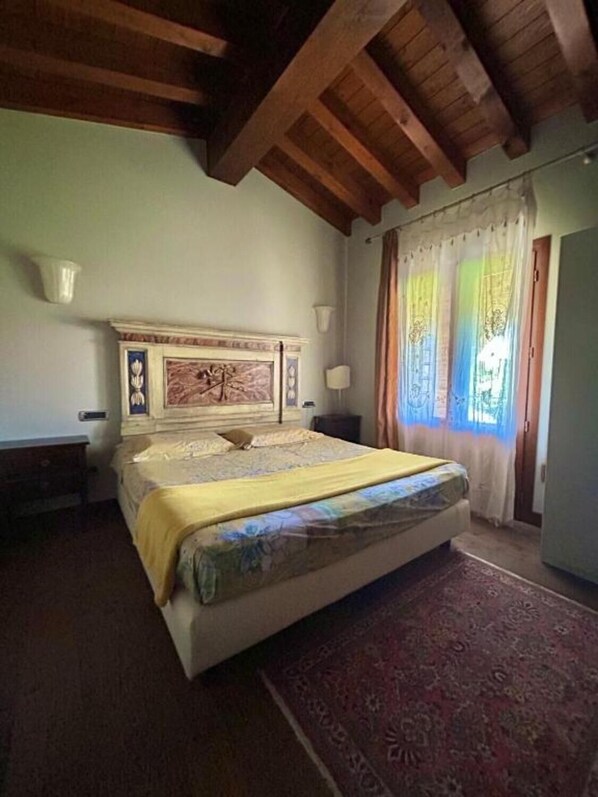 A bedroom with a large bed, wooden ceiling beams, a nightstand, a bedside table, a lamp, and a window with curtains.