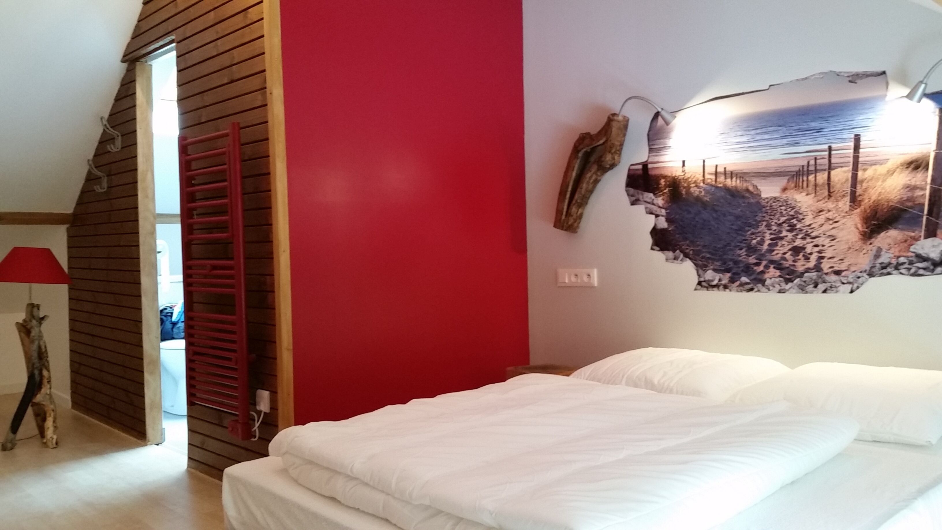 A bedroom with a red wall, a bed with white bedding, and a beach scene artwork on the wall.