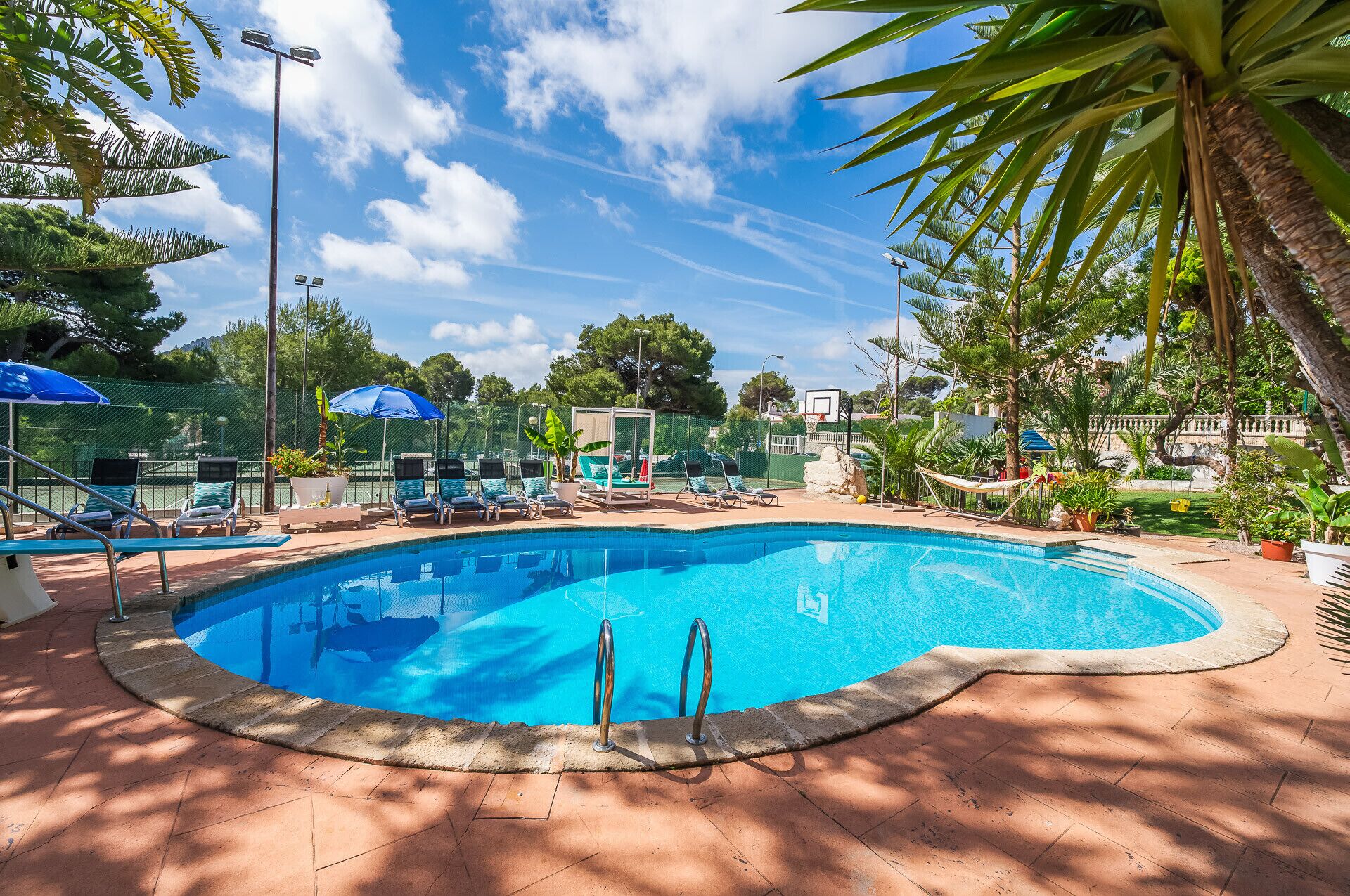 A swimming pool with lounge chairs and umbrellas, surrounded by palm trees and a basketball hoop.