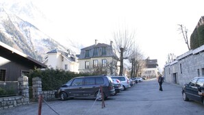 A street with parked cars and buildings, including a house with a sloped roof and a stone wall. - CHAMONIX superb stud 2/3 pers, private residence, parking, ski lift access (Chamonix-Mont-Blanc)