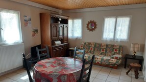 A dining area with a round table covered in a patterned tablecloth, surrounded by wooden chairs. A wooden cabinet with a decorative top is against the wall. A colorful sofa is placed against the wall, and a small table with a lamp is visible. - Les Rousses-Bois d'avont (Bois-D'amont)