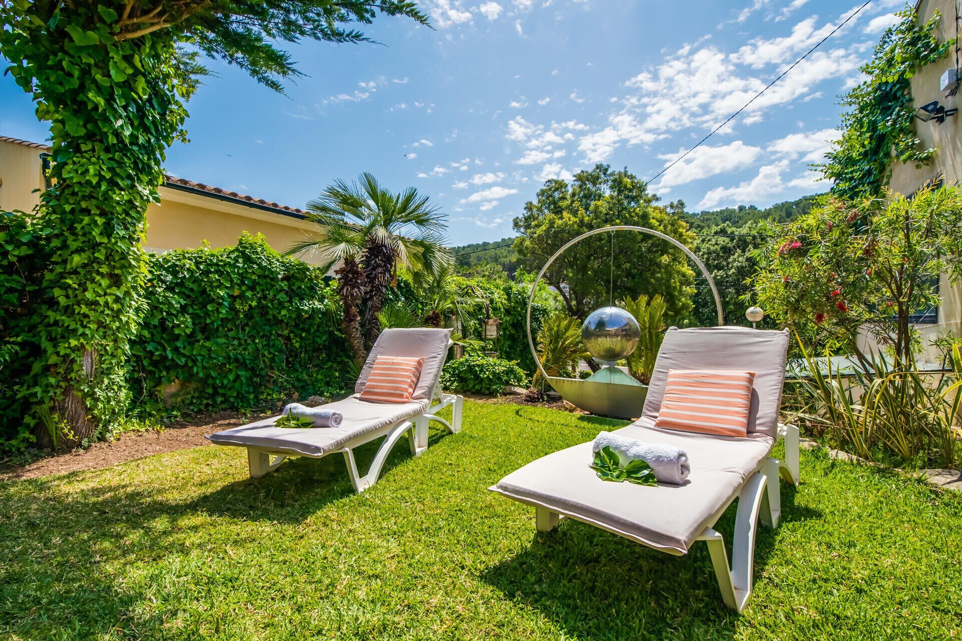 Deux fauteuils confortables avec coussins, une sphère métallique décorative et une végétation luxuriante dans un jardin.