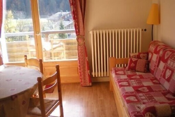 Ein gemütliches Zimmer mit Holzboden, einem rot-weißen Mustersofa, einem kleinen Tisch mit Stühlen und einem Fenster mit Blick auf einen Balkon und Gebäude.