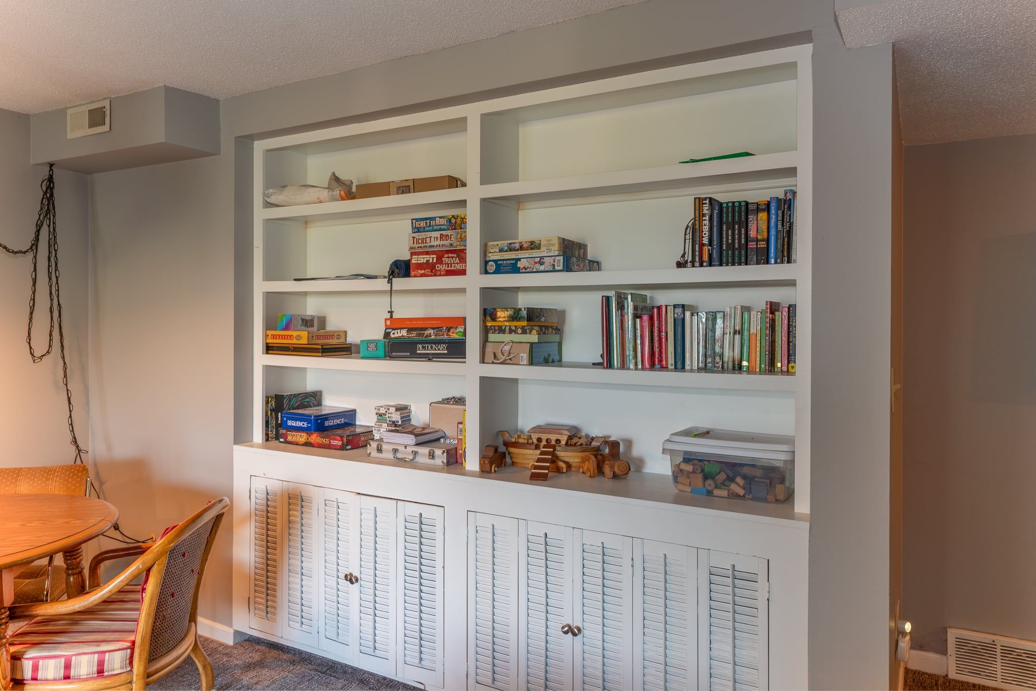 A room with a white bookshelf filled with books, games, and puzzles. There is a wooden table with chairs and a small cabinet with drawers.