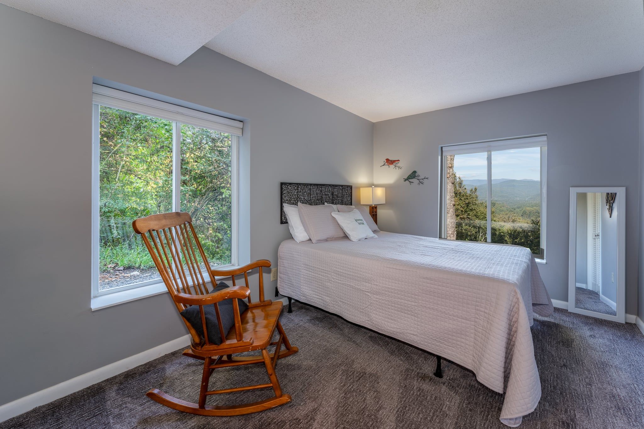 Um quarto com cama, uma cadeira de balanço, uma janela com vista e uma porta.