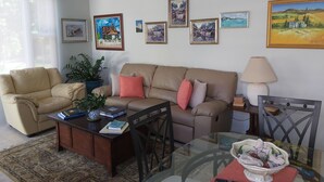 A living room with a beige sofa, a glass coffee table, and a patterned rug. There are several framed artworks on the wall and a potted plant. - Charming , Comfortable  Cottage Near Amenities (southampton)