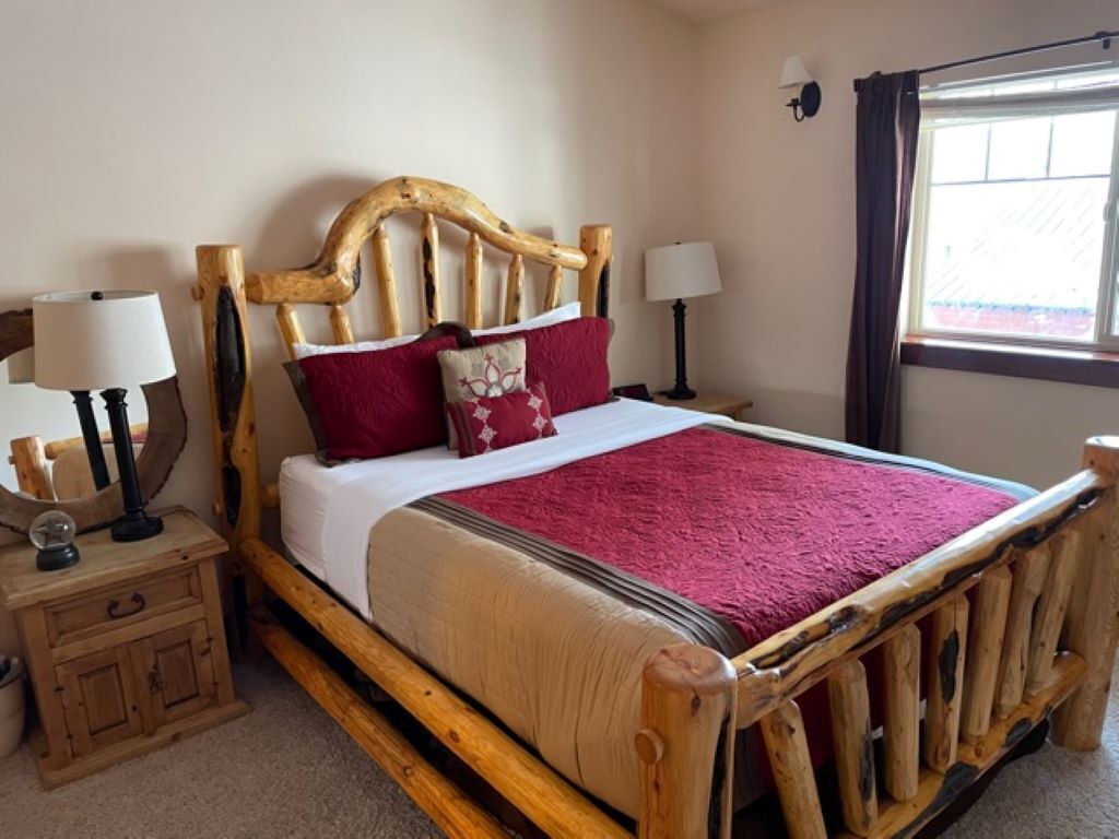 A wooden bed with a headboard, two bedside tables with lamps, a red throw blanket, and a window with curtains.