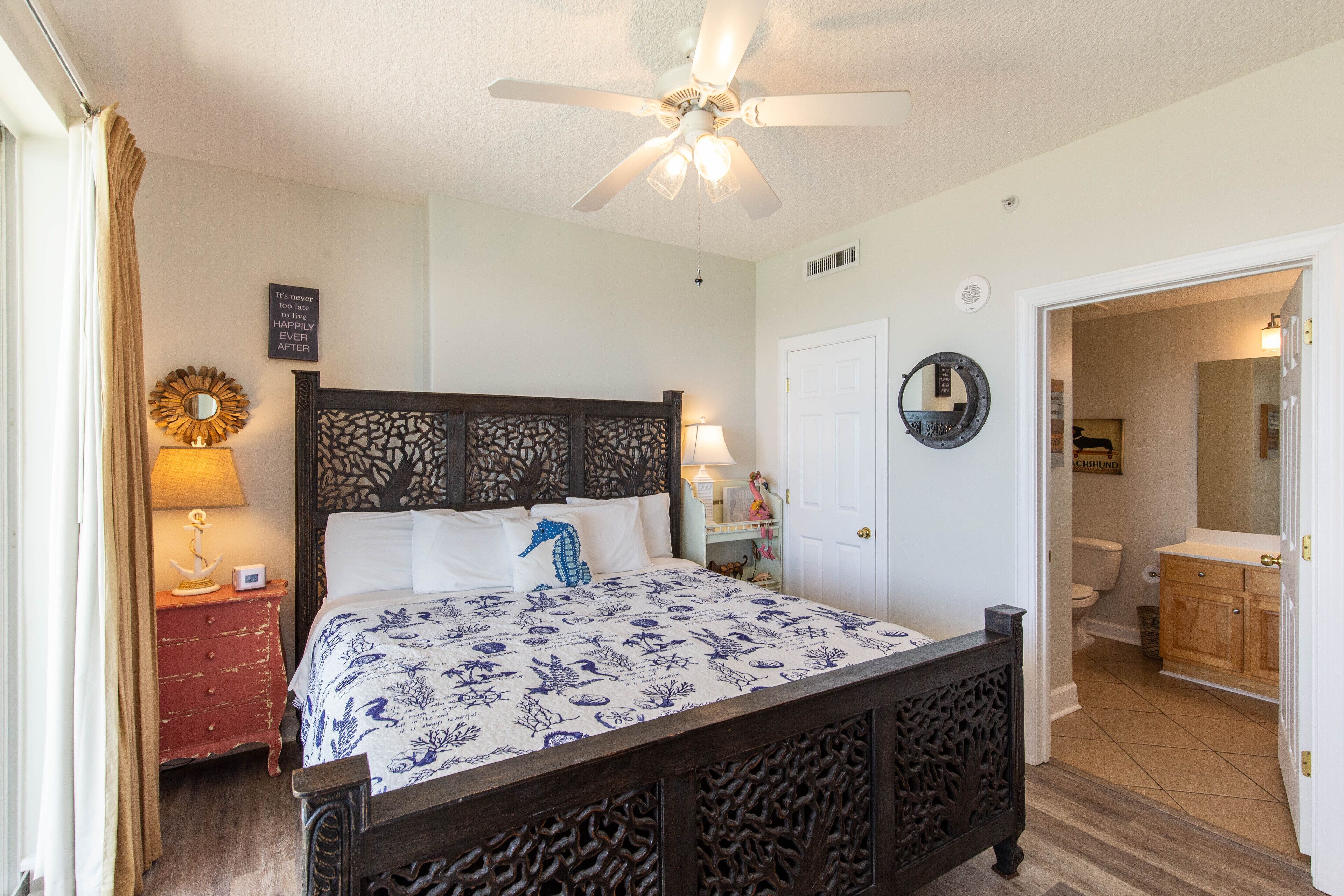 A bedroom with a bed, a ceiling fan, a nightstand, a mirror, and a door leading to a bathroom.