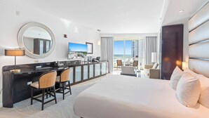 A hotel room with a large bed, a sitting area with a view, a kitchenette, and a balcony with a cityscape view.