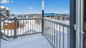 A balcony with a metal railing, snow-covered ground, and a view of a blue bus and buildings in the background. - Ski In Out Condo Walk to Village (Snowshoe)
