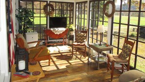 A sunlit room with a television, wicker furniture, and large windows offering a view of a green landscape.