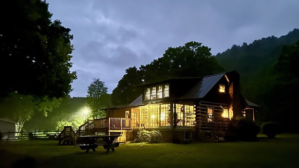 A log cabin with a porch and a picnic table in front, surrounded by trees and hills.