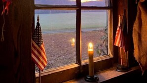 A window with a view of a sunset over a body of water, an American flag, and a lit candle on a wooden ledge.