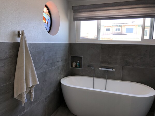 Un bagno moderno con una vasca da bagno freestanding, un porta asciugamani, una piccola finestra con vista sugli edifici e un dispenser di sapone a parete.