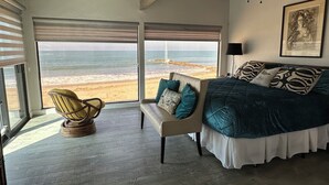 Een slaapkamer met een groot raam met uitzicht op een strand, een bed met gestreepte kussens, een stoel en een plafondventilator.