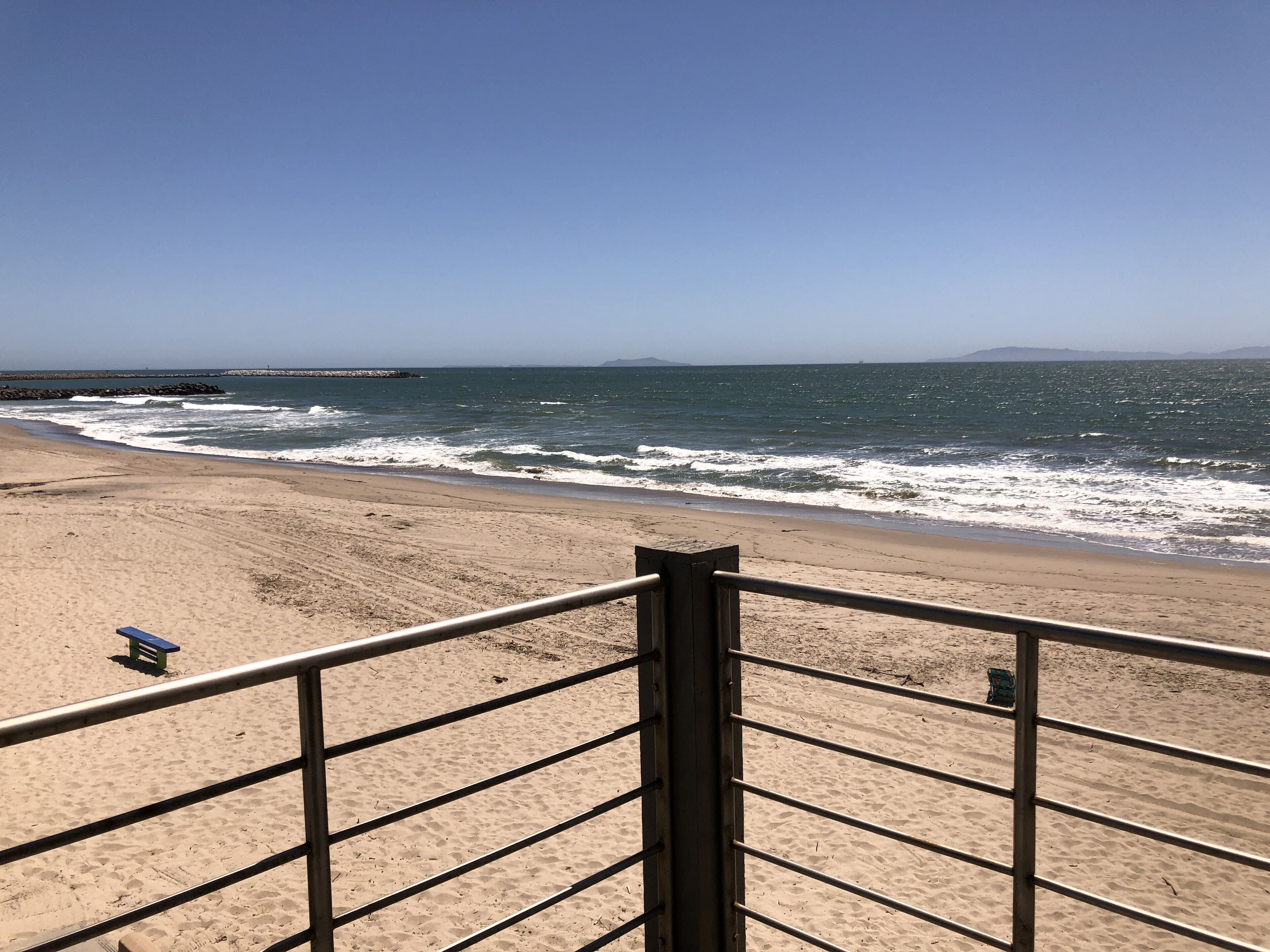 A beach with a metal railing, a bench, and a rocky breakwater in the distance.