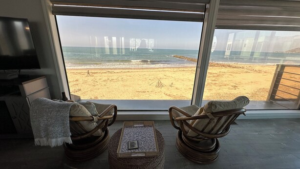 Een kamer met een groot raam met uitzicht op een strand, twee rieten stoelen, een kleine tafel en een televisie op een kast.