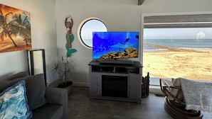 Een woonkamer met een televisie waarop een onderwaterscène te zien is, uitzicht op een strand door een raam, een bank en een potplant.