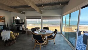 Een ruime woonkamer met een groot raam met uitzicht op een strand, een eettafel met stoelen, een televisie en een gezellige zithoek met kussens.