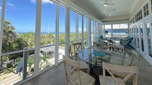 Een ruim balkon met grote ramen met uitzicht op een strand en de oceaan, ingericht met een ronde glazen tafel en houten stoelen.