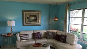 A living room with a sectional sofa, a round ottoman, a side table with a lamp, and a framed artwork on the wall. - Beautiful 1800 square foot Mid Century Home  (Venice)