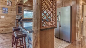 Interior de una cabaña de madera con cocina, taburetes de bar y un refrigerador.