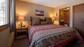 A bedroom with a bed, two bedside tables, a nightstand, a window with curtains, a painting on the wall, and a door leading to another room. - Beautifully Furnished, Clean, Cozy Cabin Next to Rocky! Walk to RMNP & YMCA (Estes Park)