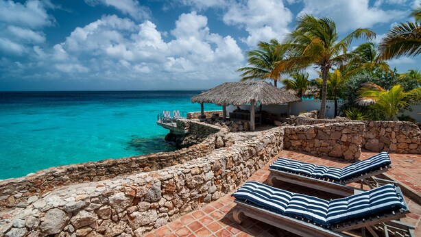 A stone-walled pool area with lounge chairs and a thatched-roof structure overlooking turquoise waters. - Ocean front Villa on the most magnificient location, only 3 steps from the ocean (Belnem Bonaire)