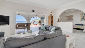 A modern living room with a grey sofa, a flat-screen TV, and a view of the beach through an arched doorway. - Ocean front Villa on the most magnificient location, only 3 steps from the ocean (Belnem Bonaire)