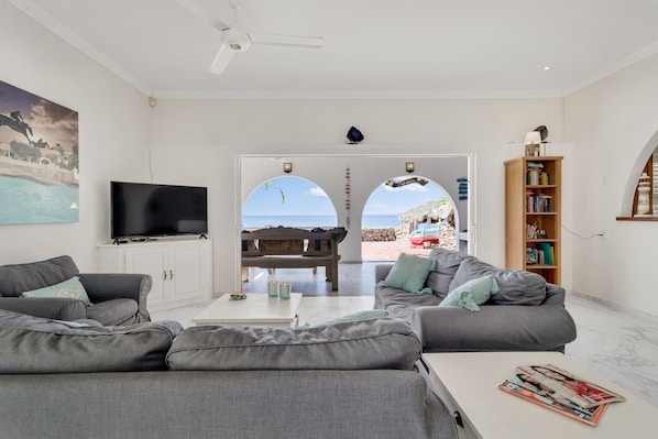 A modern living room with grey sofas, a flat-screen TV, a coffee table, and a bookshelf. There is a large window with a view of the beach. - Ocean front Villa on the most magnificient location, only 3 steps from the ocean (Belnem Bonaire)