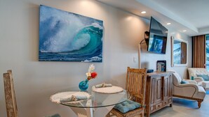A modern living room with a glass dining table, wooden chairs, a sofa, and a large wave painting on the wall. - Romantic Oceanfront Condo at Lahaina Shores with Sweeping Ocean Views (Lahaina)