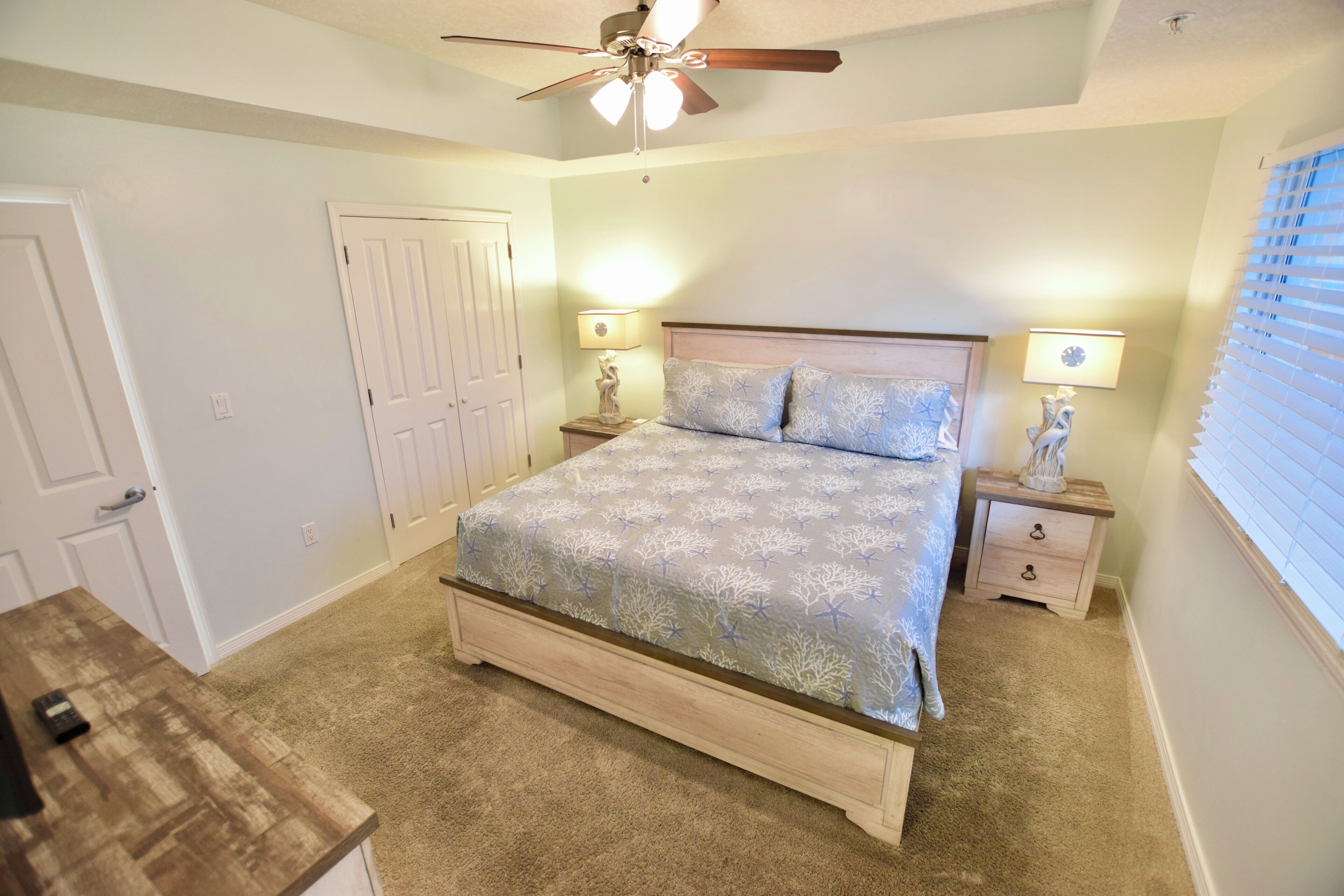 A bedroom with a bed, two bedside tables with lamps, a ceiling fan, and a door.