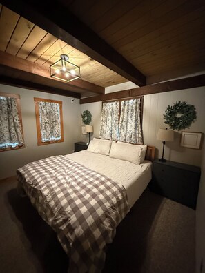 A bedroom with a large bed, wooden ceiling beams, and two windows with floral curtains. - Gore Mountain Chalet - Moments From Adventure! (Ski, Hike, Explore) (North Creek)