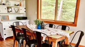 A wooden dining table with six chairs, a white tablecloth, and a window with a forest view. - Gore Mountain Chalet - Moments From Adventure! (Ski, Hike, Explore) (North Creek)