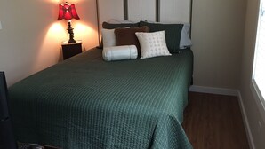 A neatly made bed with a green quilt, a bedside lamp, and a window with blinds. - The Bungalow at Brianza Winery (Crittenden)