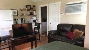 A living room with a television, a sofa, a coffee table, and a bed with a green quilt. There is a kitchen area with a microwave and a refrigerator. - The Bungalow at Brianza Winery (Crittenden)