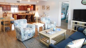 A living room with a sofa, chairs, a coffee table, a TV, and a kitchen in the background. - Beautiful and Spacious, 2 bed 2 full bath, pool and jacuzzi, steps to the ocean (Hatteras)