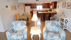 A living room with two patterned armchairs, a small white table with a lamp, a dining area with a wooden table and chairs, and a kitchen in the background. - Beautiful and Spacious, 2 bed 2 full bath, pool and jacuzzi, steps to the ocean (Hatteras)