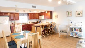 A compact kitchen with wooden cabinets, a central island with bar stools, a dining table with chairs, and a bookshelf. - Beautiful and Spacious, 2 bed 2 full bath, pool and jacuzzi, steps to the ocean (Hatteras)