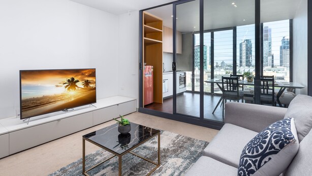 A modern living room with a large TV, a sofa, a coffee table, and a view of the city through large windows. - CityLights apartments (Melbourne)