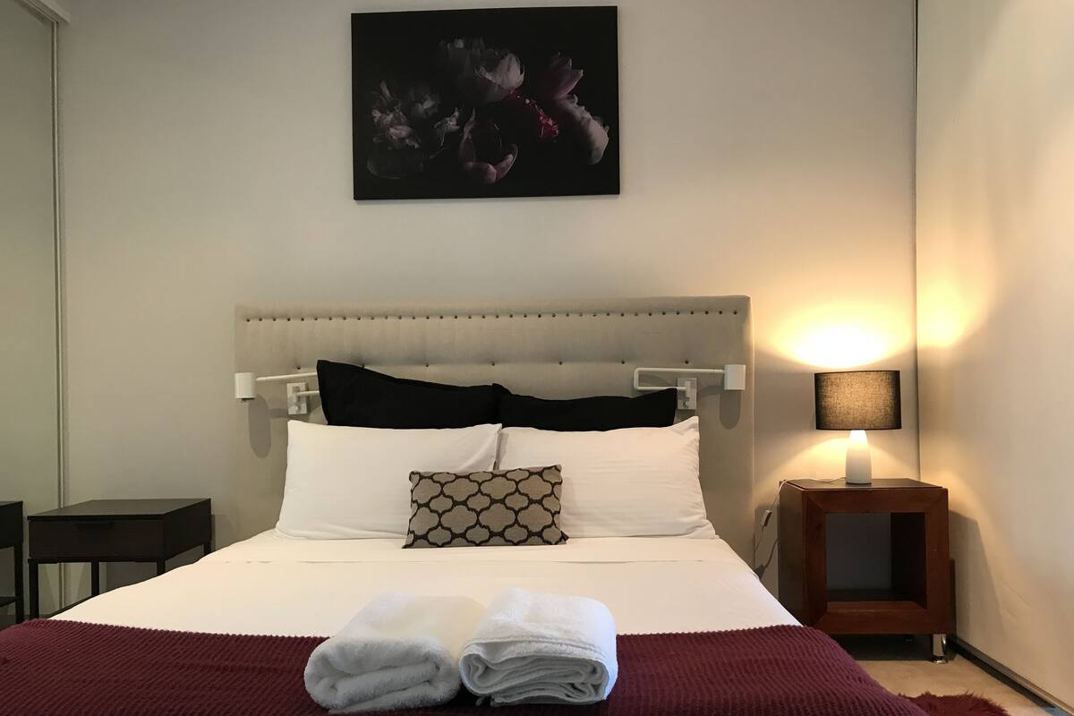 A neatly made bed with white linens and a folded towel, a bedside table with a lamp, and a wall-mounted artwork above the bed.
