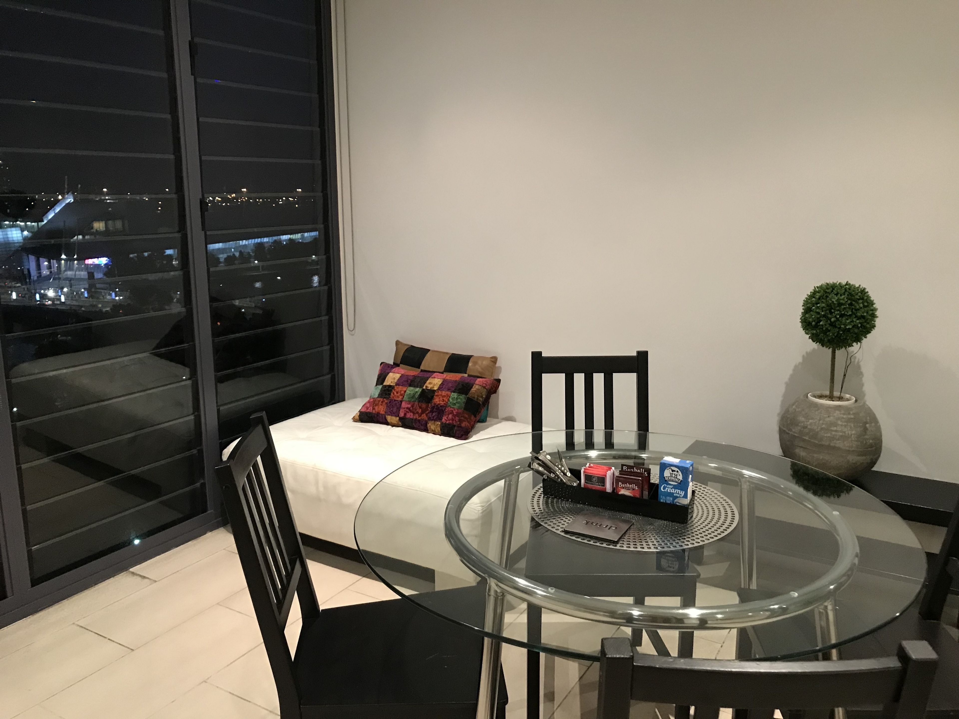 A modern room with a glass table, black chairs, a small sofa with a colorful pillow, a potted plant, and a view of the city at night.