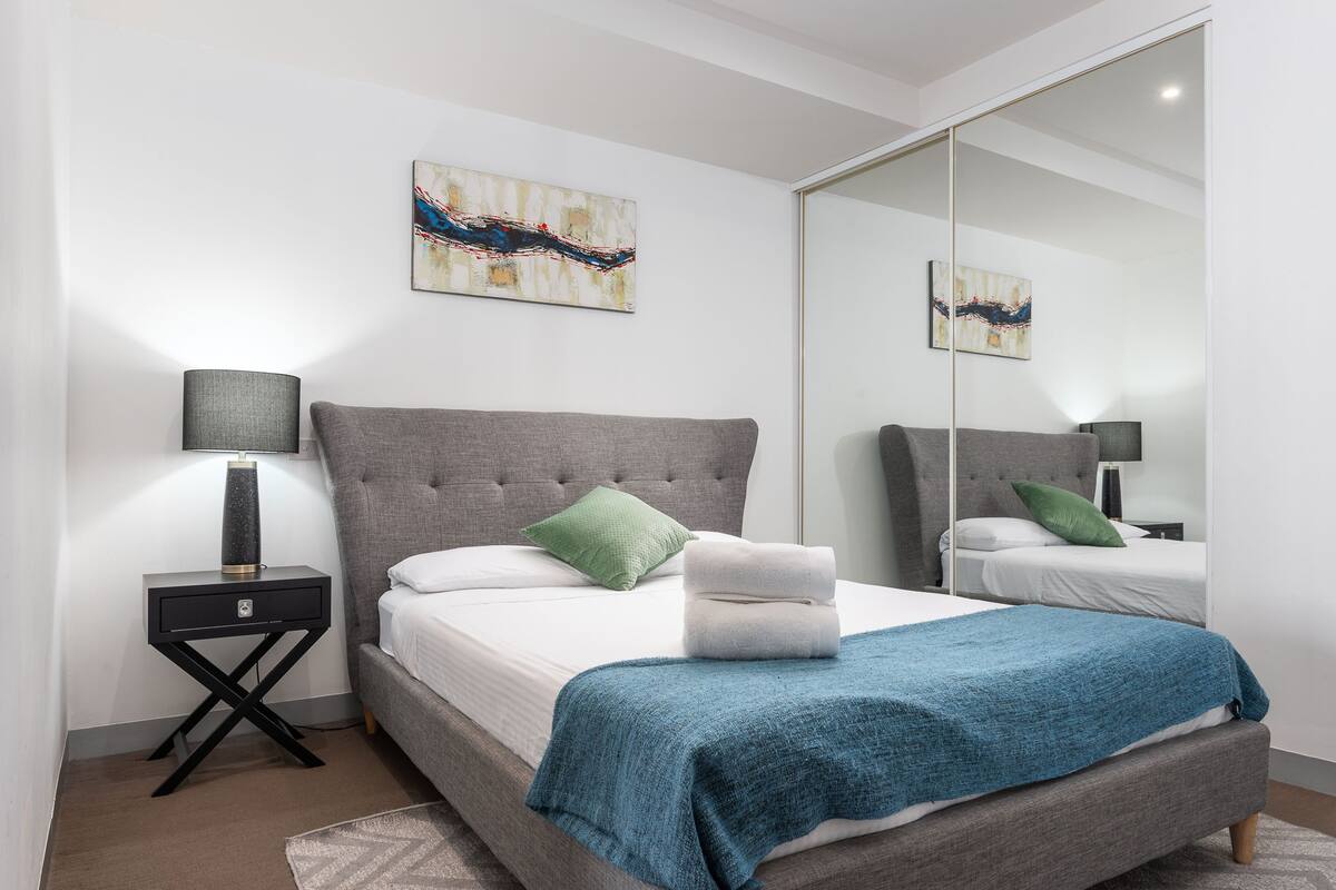 A modern bedroom with a grey upholstered bed, a blue throw blanket, a bedside lamp, and abstract artwork on the walls.