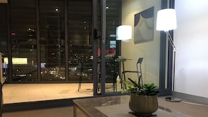 A modern hotel room with a large window offering a city view, a small table with a potted plant, and a floor lamp. - CityLights apartments (Melbourne)