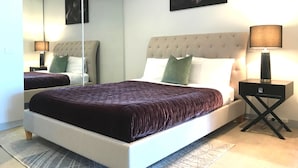 A bedroom with a large bed, a nightstand, a lamp, a mirror, and a painting on the wall. - CityLights apartments (Melbourne)